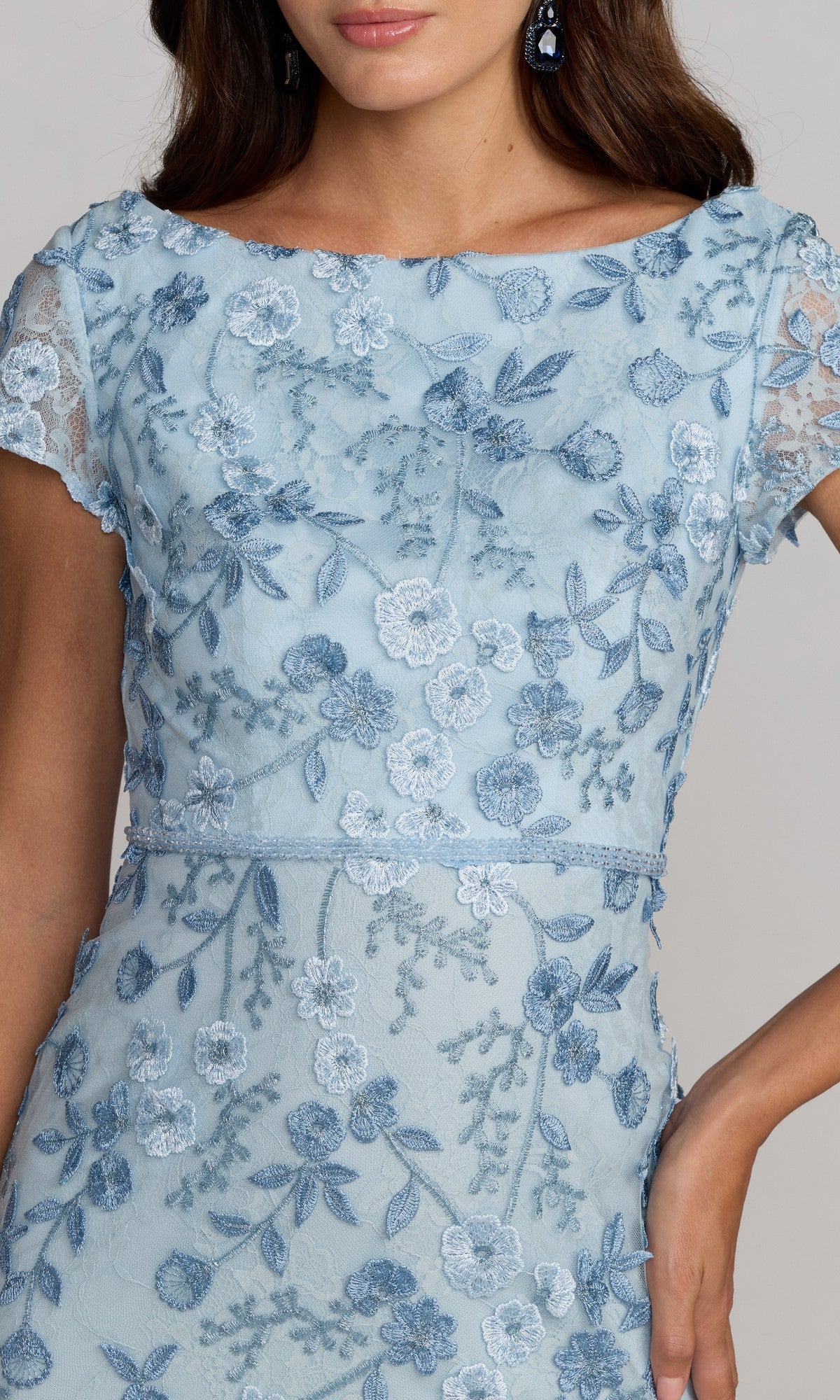 A woman wears the Long Formal Dress 67711 by Mac Duggal, a light blue lace gown with floral embroidery and short sleeves. She accessorizes with matching earrings and rests her right hand on her hip.